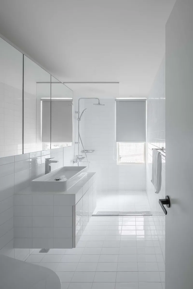 White-tiled bathroom with floating vanity, glass shower partition and windows with translucent blinds