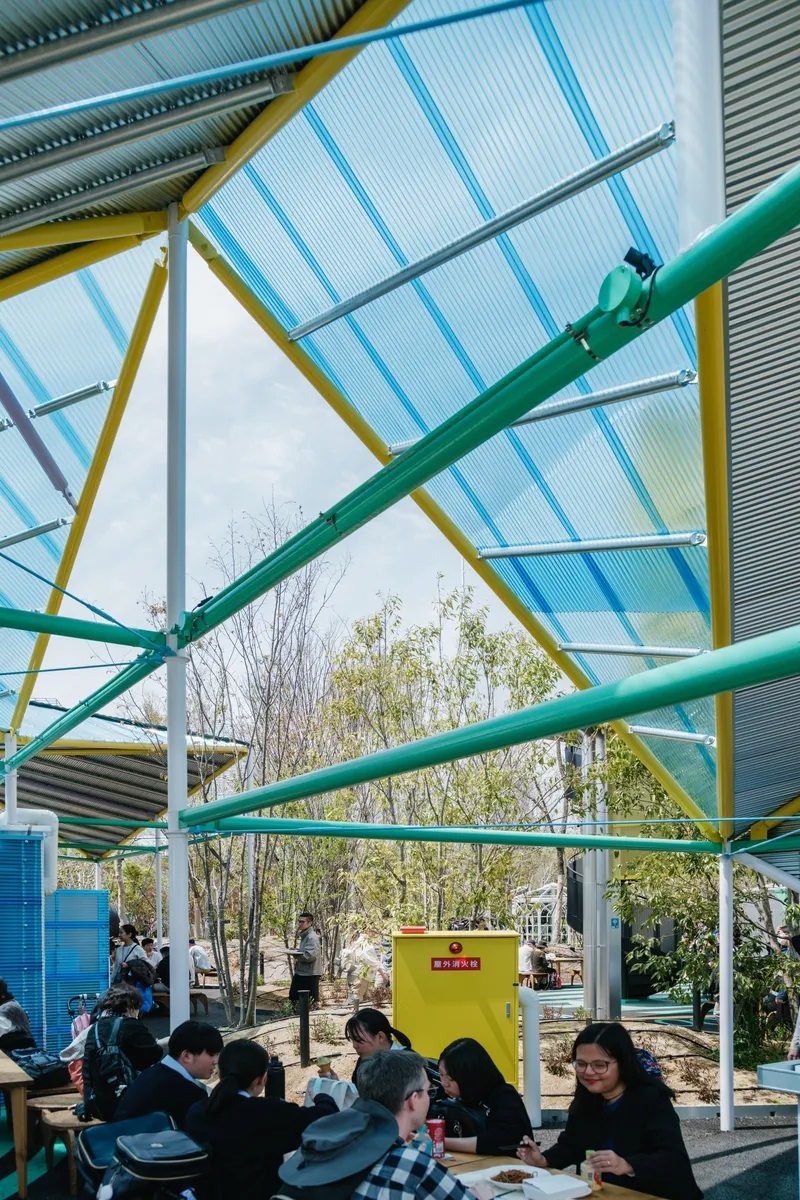 Underside of the canopy showing yellow and green painted steel trusses with translucent corrugated roofing panels