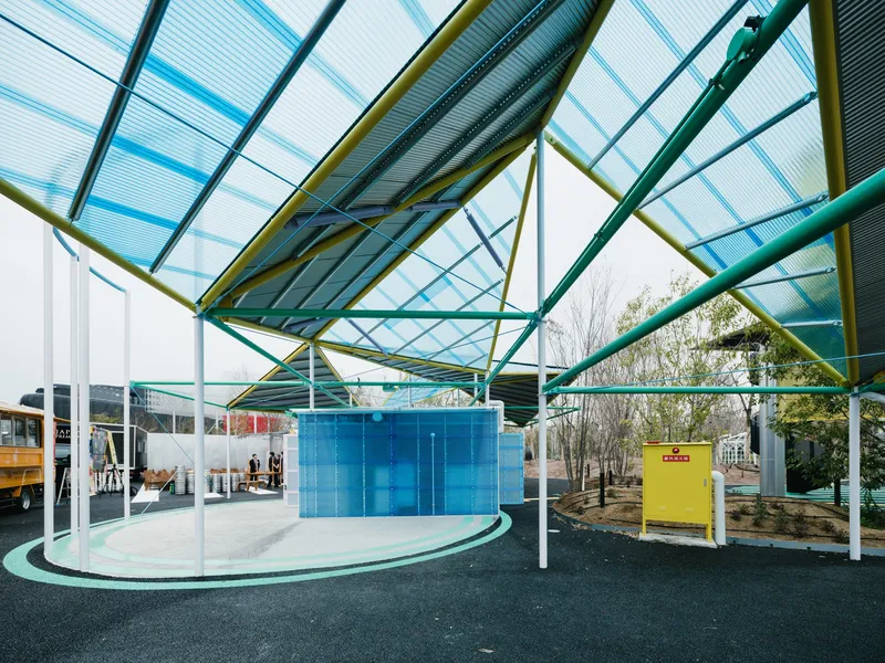 Covered outdoor plaza with colored steel columns and translucent corrugated canopy over asphalt ground with painted graphics