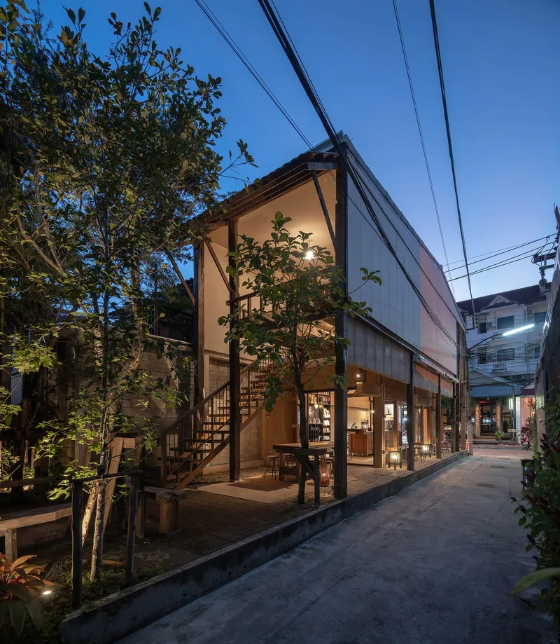 Street view of the translucent polycarbonate and timber facade illuminated at dusk beside potted trees