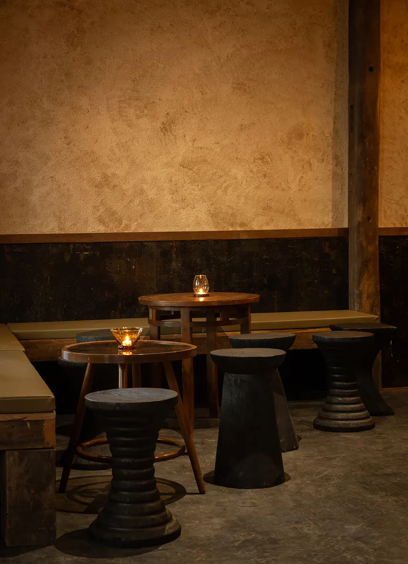 Low timber tables and turned stools against a two-tone plaster wall lit by candlelight