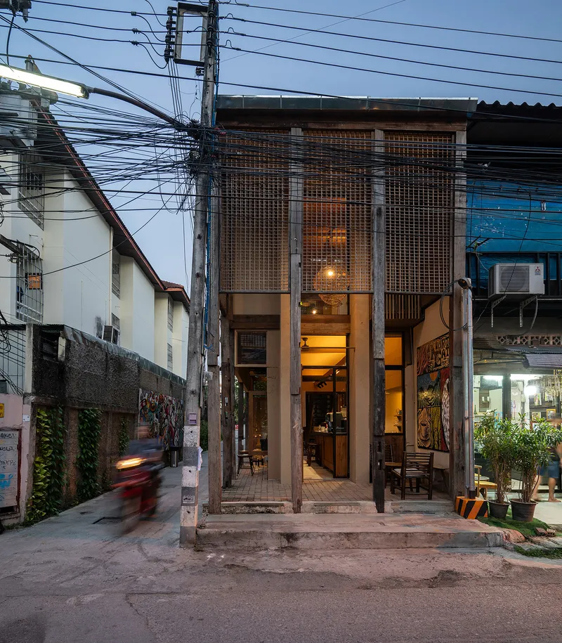 Narrow street facade with perforated metal screen panels, exposed columns and overhead power lines at twilight