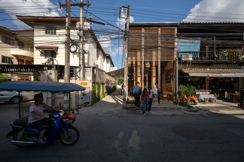 Street view of a metal screen facade with pedestrians and a motorcyclist passing beneath overhead wires