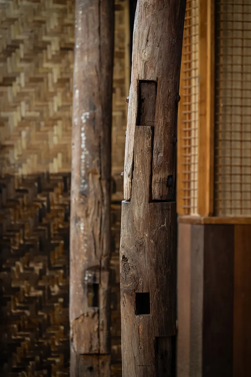Close-up of mortise and tenon joinery in a weathered timber column against woven bamboo wall panels