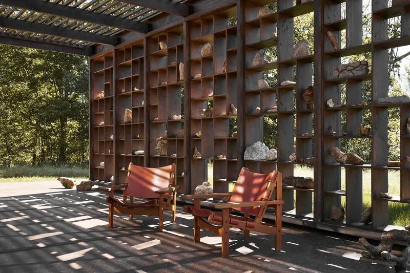 Open-air terrace with timber pergola, rock-filled shelving grid, and leather lounge chairs in dappled shade