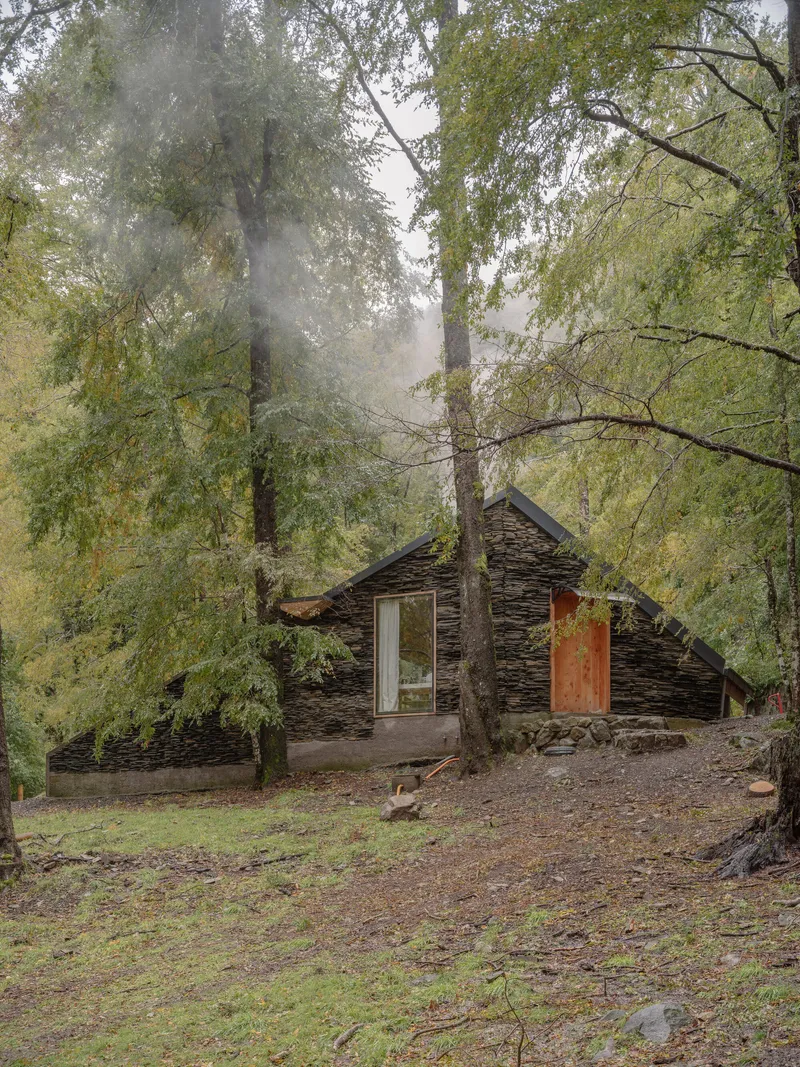 Gabled structure with stacked slate facade and timber door emerging from a misty forested hillside