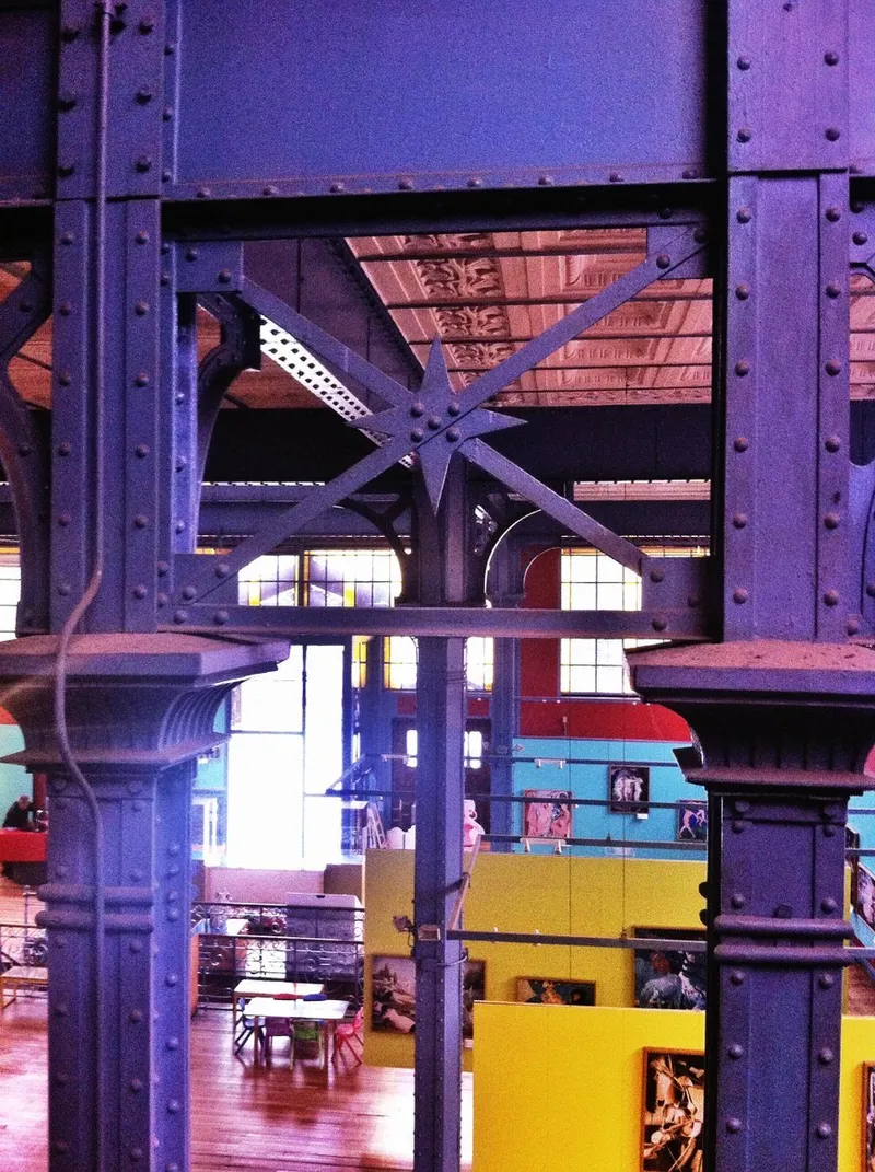 View through the riveted steel trusses at dusk with industrial ceiling and colored exhibition spaces visible beyond