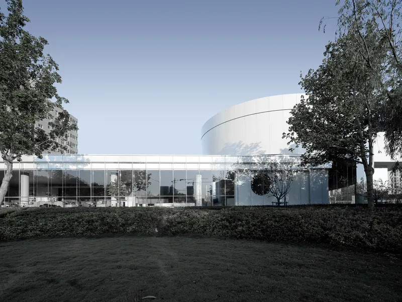 Glass-walled pavilion connecting to cylindrical volume with mature trees framing the composition