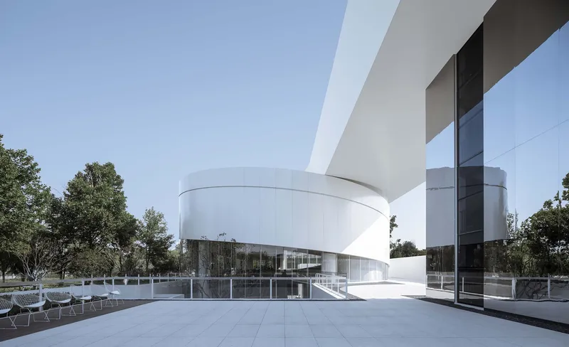 White cantilevered bridge connecting cylindrical volume to glass pavilion with pool and trees beyond