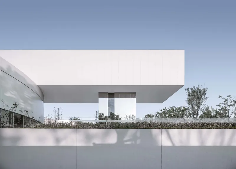 Cantilevered upper volume above courtyard with planted beds and young trees in afternoon light
