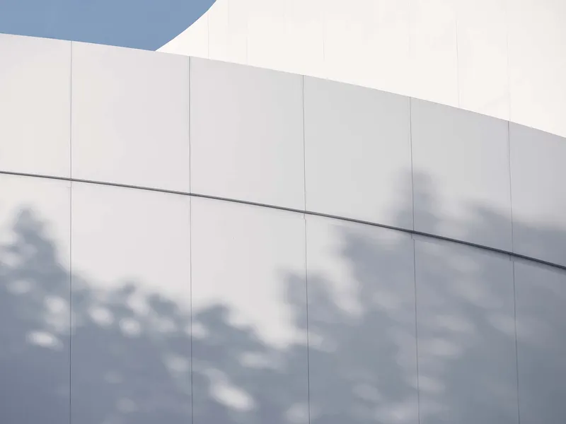 White panel facade with horizontal reveals casting blurred tree shadows across the surface
