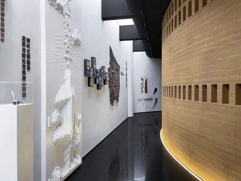 Gallery corridor with white walls featuring relief maps and exhibits alongside a curved timber-clad volume with uplighting