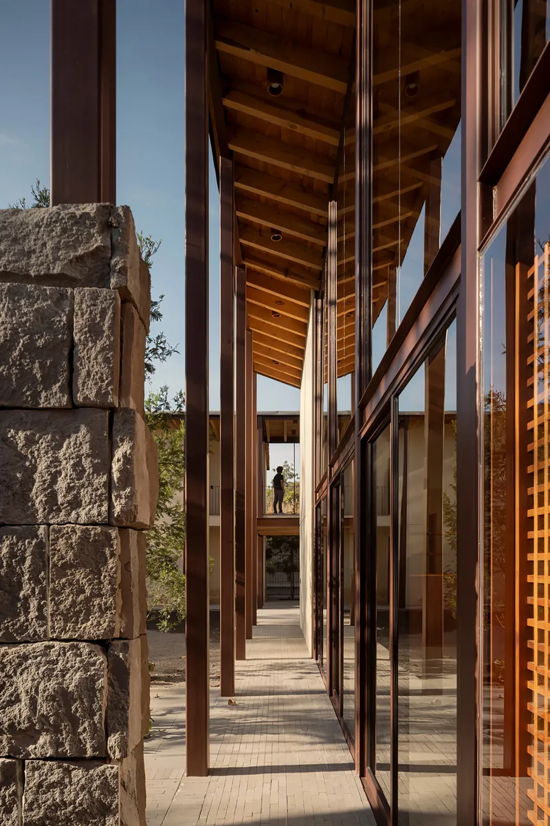 Narrow glazed corridor framed by steel columns and a rough-hewn stone wall