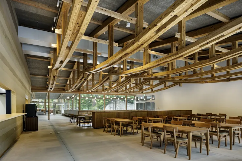 Open dining area with intersecting timber trusses overhead, concrete ceiling inserts, and rows of wooden tables and chairs