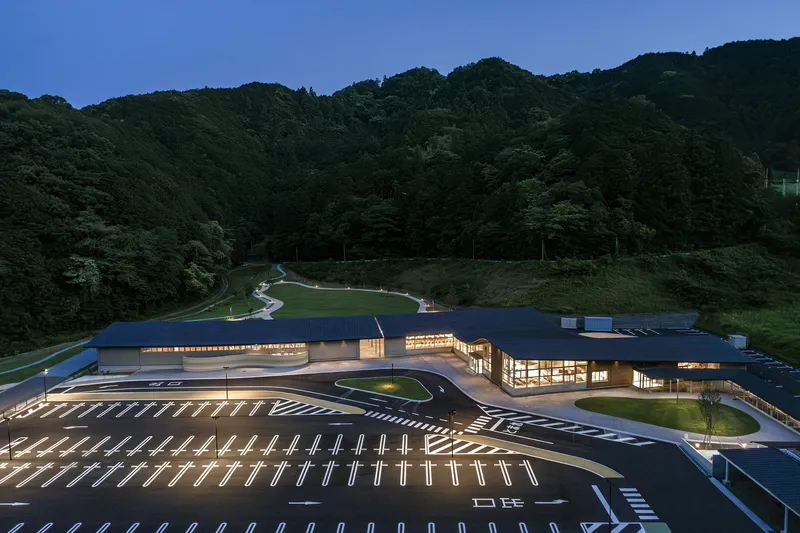 Illuminated glass facade and parking lot at twilight with forested hills rising behind
