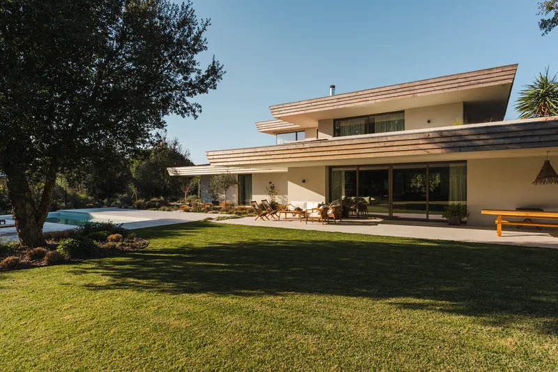 Rear elevation showing layered roof overhangs and glass walls opening to terrace and lawn in morning light