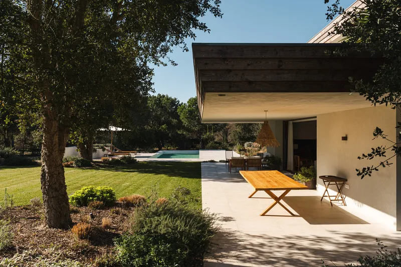 Covered outdoor dining area with timber ceiling extending to garden and pool beneath deciduous trees