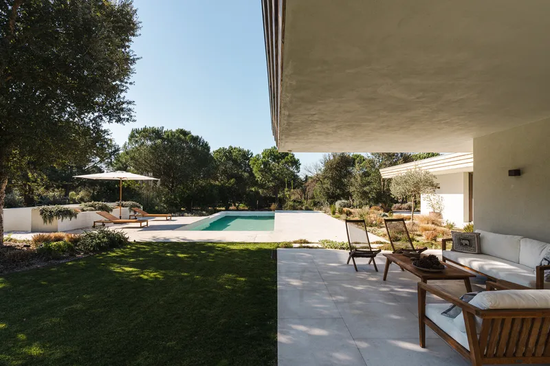 Covered terrace with wooden lounge chairs overlooking the pool and lawn in dappled sunlight