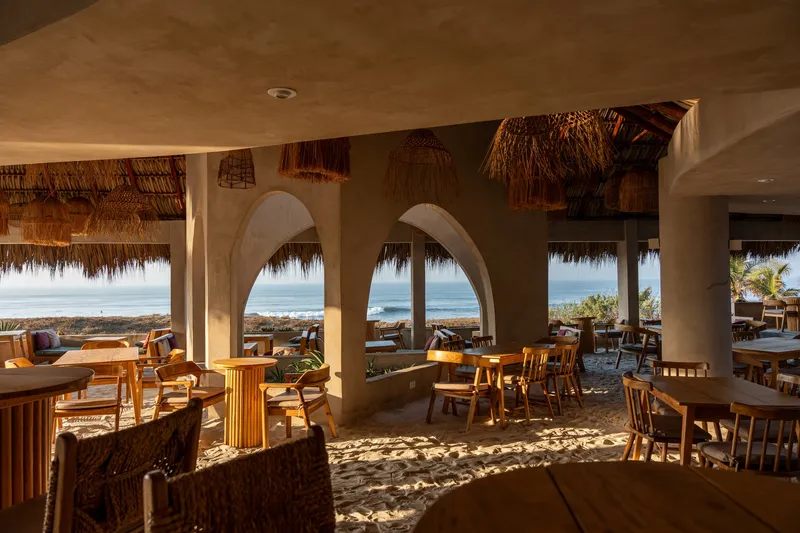 Dining area with earthen arches framing ocean views and dappled sunlight across sand floor