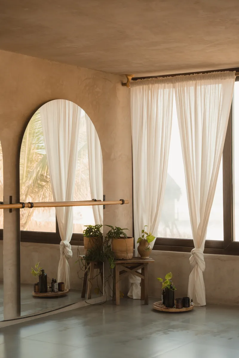Arched mirror with timber shelf and potted plants against plaster wall with sheer curtains filtering daylight