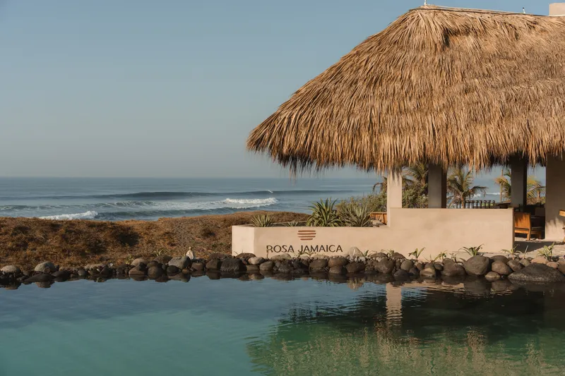 Thatched pavilion and stucco wall beside a turquoise pool with rock edging and ocean beyond