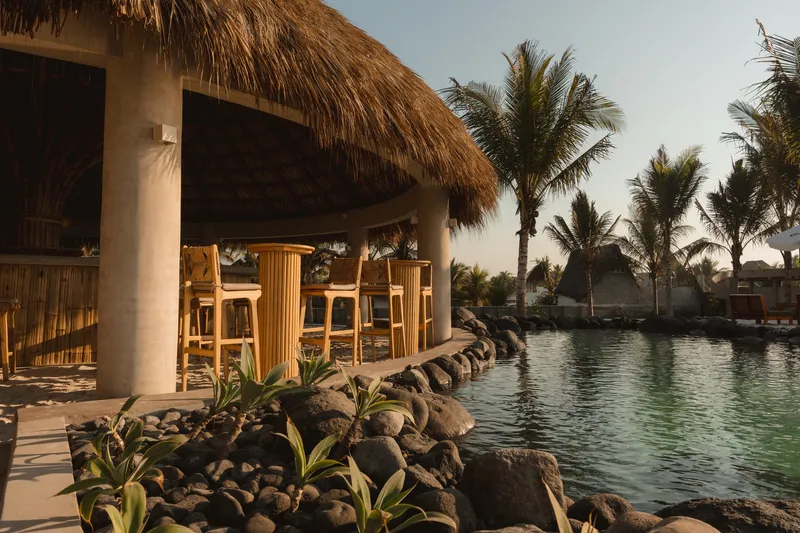 Poolside bar pavilion with thatched roof, concrete columns and timber stools beside a rock-edged lagoon at dusk