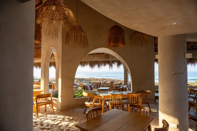 Dining terrace with arched openings framing ocean views, woven pendant lights and dappled sunlight on the floor