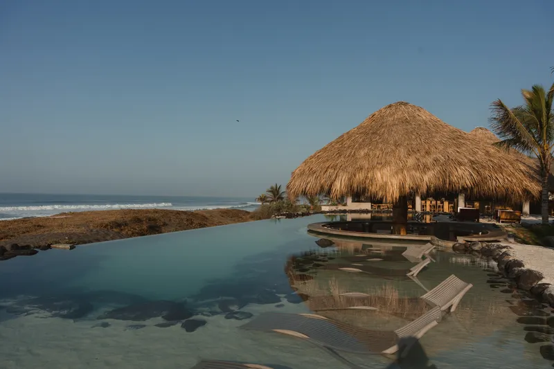 Infinity pool edge with submerged loungers and thatched pavilion against the ocean at dawn
