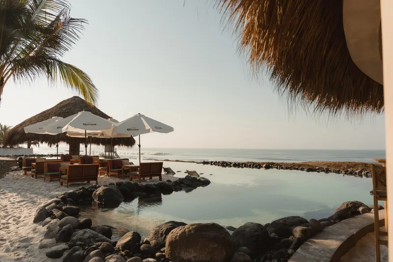 Rock-edged saltwater pool with thatch-roofed pavilion and lounge seating near the ocean at dusk