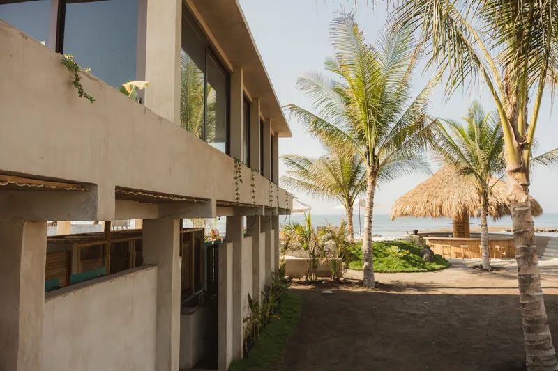 Two-story stucco volume with horizontal windows beside palm trees and thatch-roofed structures near the beach