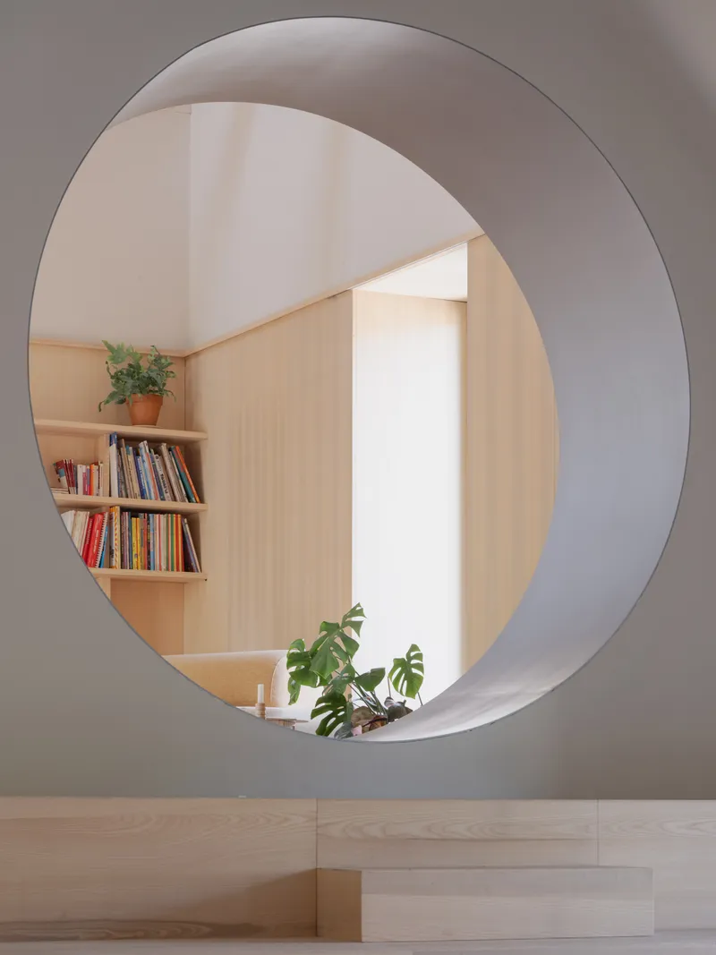 Circular portal opening framing plywood shelving with books and potted plants in adjacent room