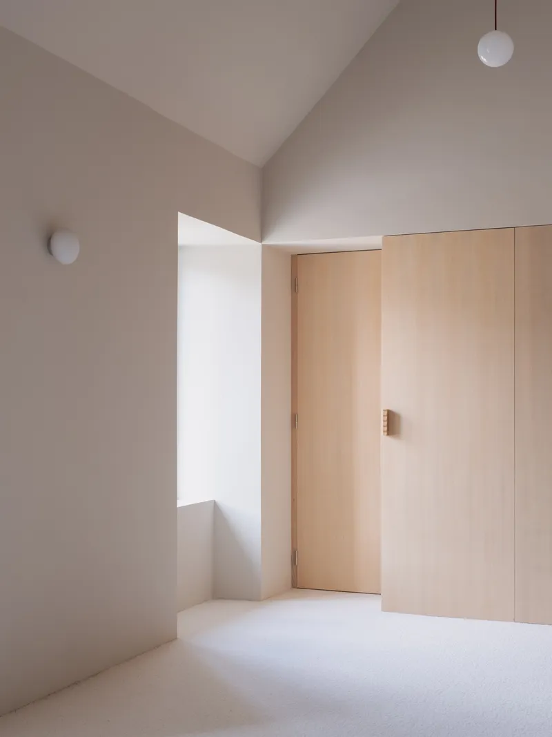 Vaulted bedroom interior with plywood wardrobe doors and round wall sconce in pale plaster walls