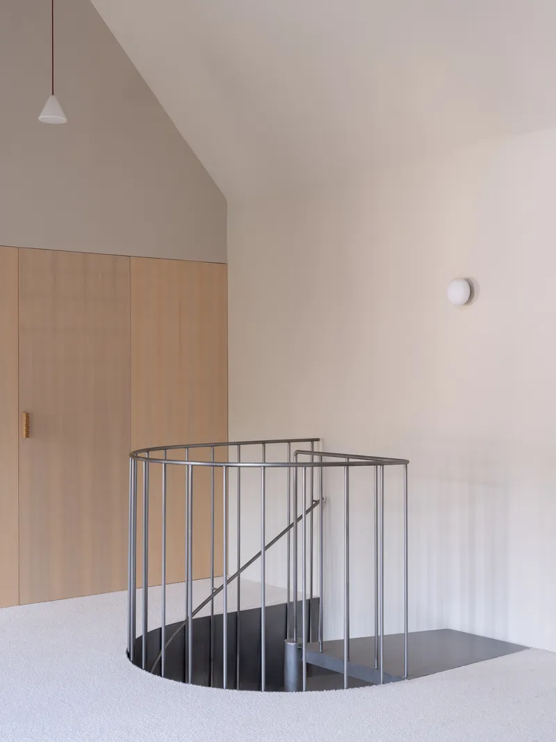 Steel spiral staircase opening framed by plywood wardrobe and white plaster walls under pendant light