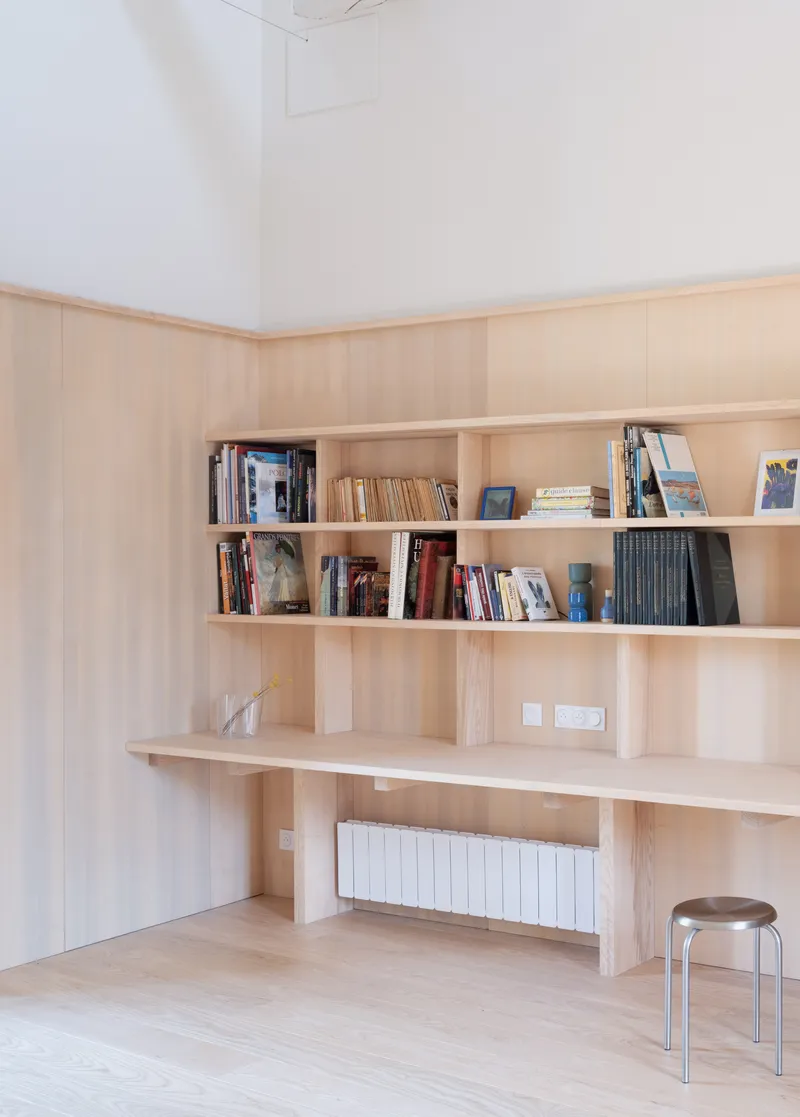Corner built-in pale timber shelving with integrated desk and radiator beneath a white vaulted ceiling