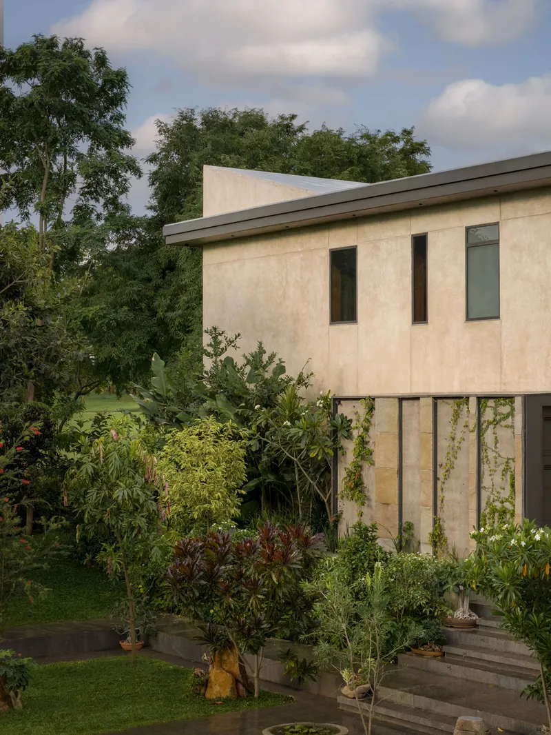 Cream-rendered facade on pilotis with narrow vertical windows and lush garden planting below