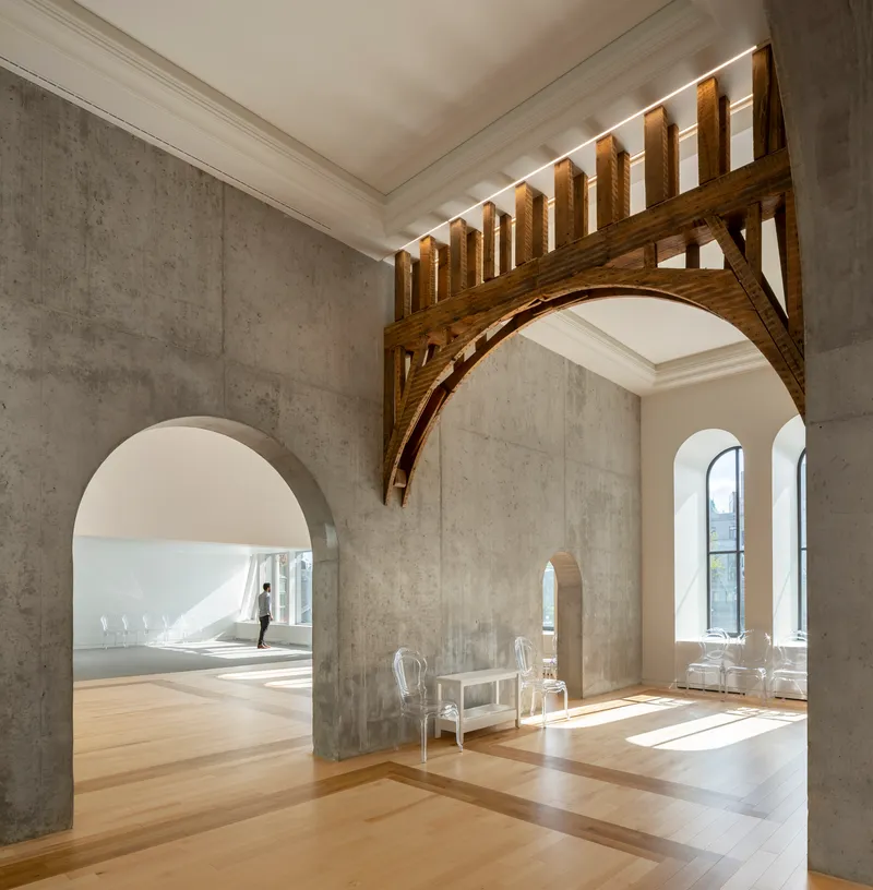 Concrete wall with arched opening and timber truss support beneath a slatted wooden screen in bright sunlight