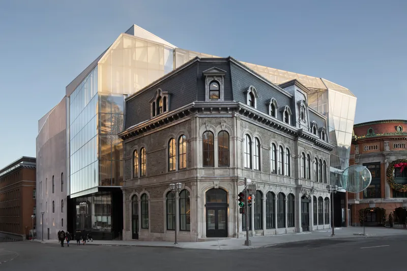 Street corner view of the historic masonry facade wrapped by a translucent glass volume at dusk