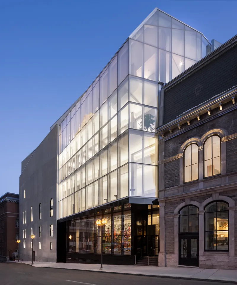 Side elevation showing the illuminated translucent glass facade rising behind the historic brick facade at dusk