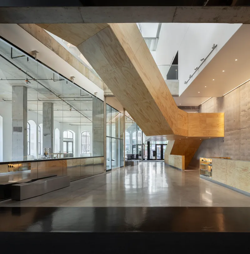 Entrance lobby with angular plywood-clad staircase beside glass-enclosed gallery and polished concrete floor
