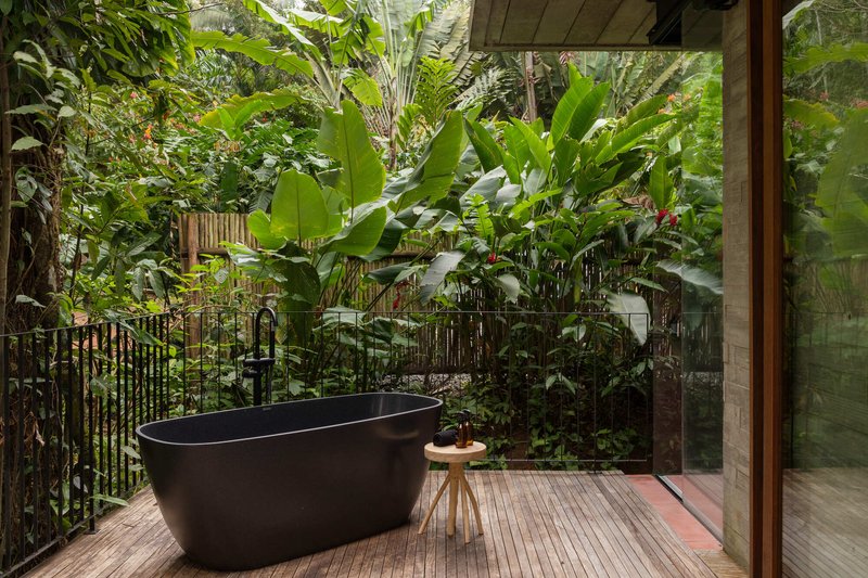 Black freestanding bathtub on timber decking with banana plants and glass sliding doors behind