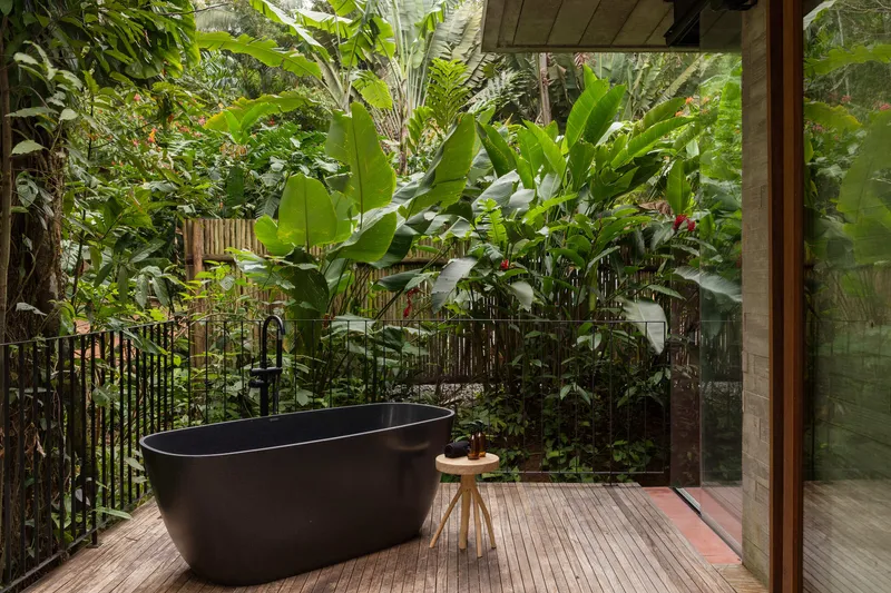 Black freestanding bathtub on timber decking with banana plants and glass sliding doors behind