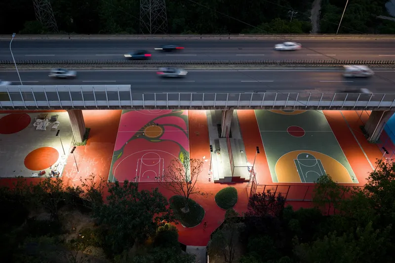 Illuminated basketball courts in pink and orange beneath a highway with evening traffic overhead