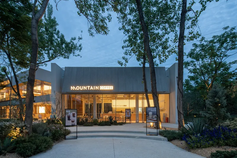 Coffee shop facade with corrugated metal cladding and glazed storefront framed by mature trees at twilight