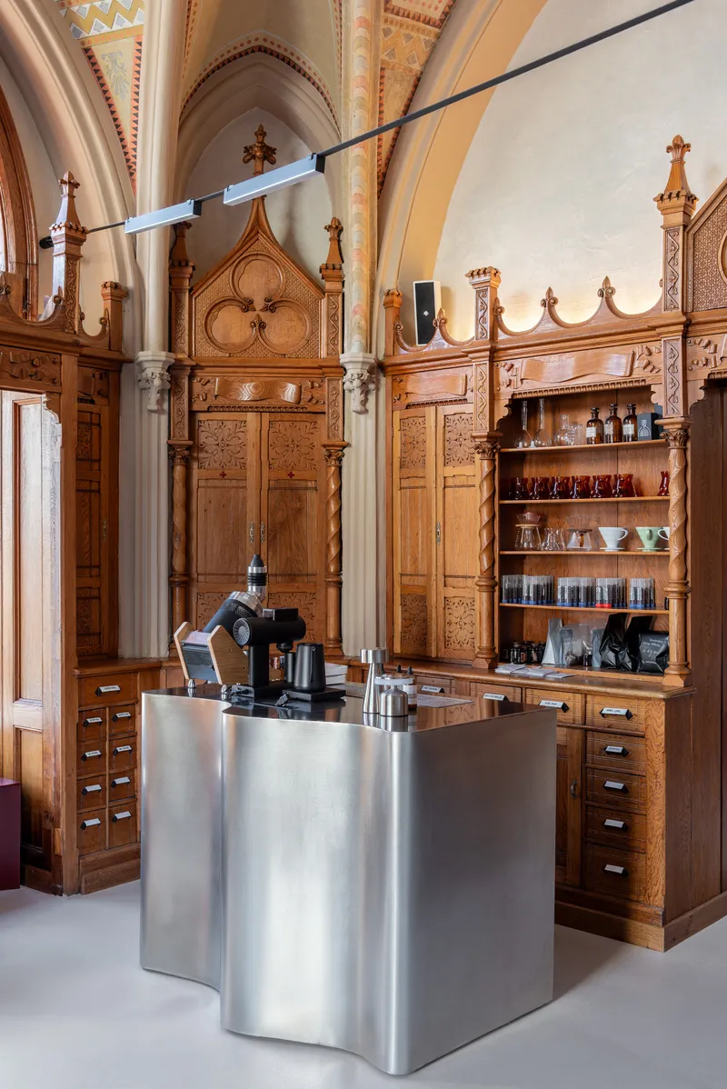 Single undulating stainless steel counter with coffee equipment below pointed arch cabinetry and painted vaults