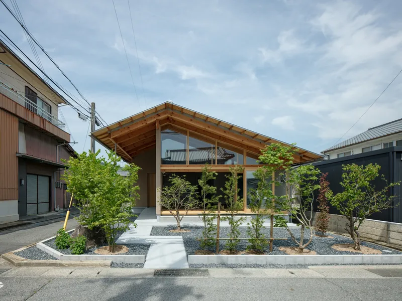 Street view of the timber-framed gable facade with glazing and gravel garden planted with young trees