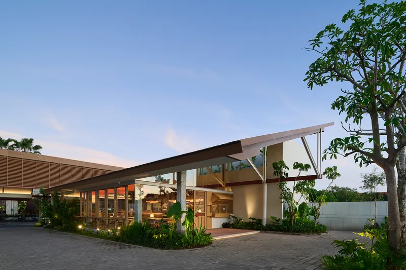 Entry facade with angled canopy structure and glass walls framed by tropical landscaping at twilight
