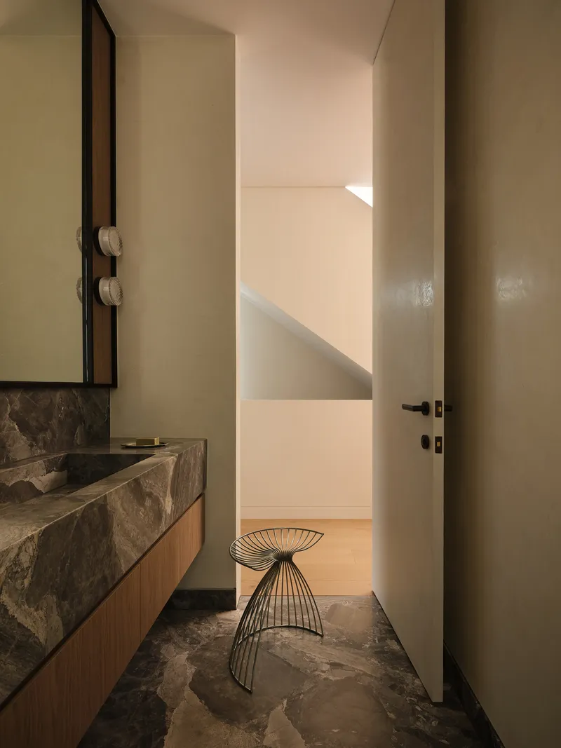 Powder room with stone vanity, wire stool, and view through open door to staircase beyond