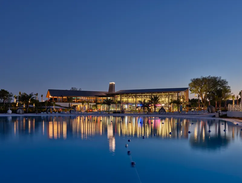 Glazed pavilion reflected in a pool at blue hour with fountain jets and palm trees