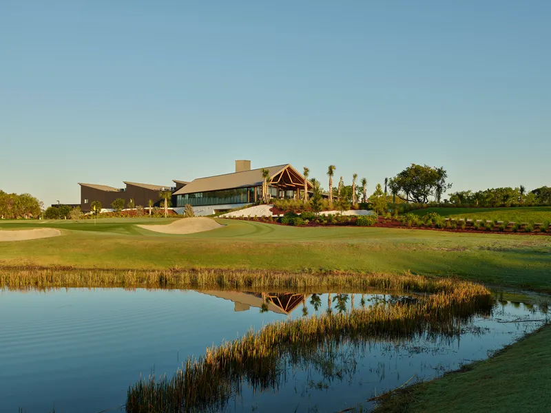 Clubhouse complex viewed across golf course water hazard with reedy shoreline in late afternoon light
