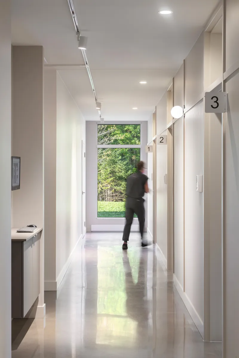 White corridor with polished concrete floor and figure in motion passing a large window to green trees